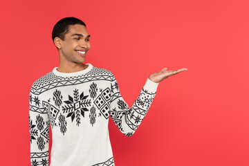 Wall Mural - smiling african american man in knitted winter sweater with ornament pointing with hand isolated on red