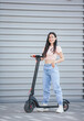 A young beautiful hipster woman is standing next to her electric scooter with modern architecture in the background.