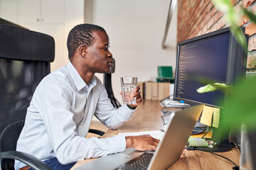 Programmer drinking water while reviewing code on computer while working in small office