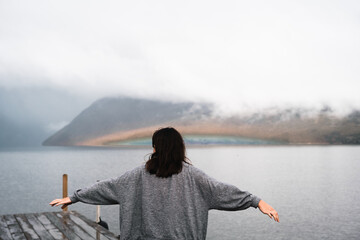 Canvas Print - caucasian girl with gray shirt and loose hair on her back with arms outstretched relaxed and calm contemplating the beauty of the calm lake near the mountains, nelson lakes, new zealand