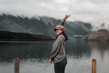 Canvas Print - caucasian young woman standing happy and content in gray shirt and brown beanie on her head smiling and pointing to the sky with finger and arm outstretched upwards at calm lake near mountains, nelson