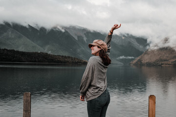 Canvas Print - caucasian girl standing relaxed and happy smiling near lake wearing brown hat gray shirt and blue jeans with right hand stretched upwards with mountains in the background on a foggy day, nelson lakes