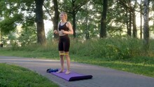 Slender Athletic Girl In Black Tight-fitting Tracksuit Does Squats With Elastic Band On Street In Park On Purple Fitness Mat. Young Woman Goes In For Sports In Morning Outside Sports Ground.