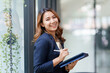 © amnaj - Attractive smiling Asian businesswoman standing holding tablet working and recording work details in office.
