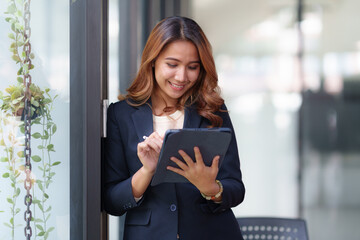 Attractive smiling Asian businesswoman standing holding tablet working and recording work details in office.