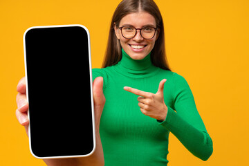 Wall Mural - Happy young woman pointing to blank phone screen, isolated on yellow background