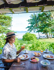Wall Mural - women with a breakfast table on the beach with palm trees in Thailand. colorful breakfast with eggs and fruit with a look at the ocean from the restaurant