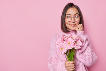 Wall Mural - Attractive dark haired Asian woman looks aside thoughtfully has romantic mood wears transparent eyeglasses jumper holds beautiful bouquet of gerbera flowers isolated over pink studio background