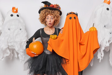 Wall Mural - Impressed shocked woman with curly hair wears black dress and hat holds pumpkin poses near orange ghost stares amazed at camera believes in magic celebrates Hallooween comes on costume party