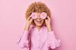 Happy curly haired young woman covers eyes with gerbera flowers smiles gladfully has fun dressed in fashionable blouse isolated over pink background. People florist and positive emotions concept