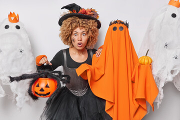 Wall Mural - Frightened female witch holds carved pumpkin with big scary spider wears black hat and dress stands near spooky ghost celebrates Halloween enjoys party time. October celebration and holidays concept