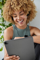 Wall Mural - Photo of cheerful curly haired woman concentrated in digital tablet connected to headphones watches funny movie during spare time dressed in t shirt poses indoors. People and technology concept