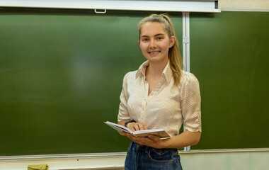 Wall Mural - A young woman with a book at the chalkboard smiles. A young teacher at school.