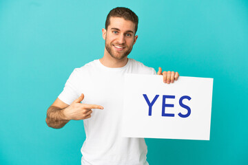 Wall Mural - Handsome blonde man over isolated blue background holding a placard with text YES and  pointing it