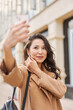 © olga pink - Glamour attractive asian kazakh female wearing beige coat, taking selfie, checking haircut, look at smartphone camera, walking on street
