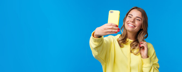 Close-up portrait tender cute blond girl in yellow hoodie, holding mobile phone, taking selfie with smartphone posing adding photo filter in appllication, standing blue background