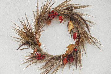 Stylish autumn wreath hanging on white wall outdoor. Fall decor and arrangement of farmhouse. Thanksgiving. Rustic autumn wreath with dried grass, berries, herbs and leaves