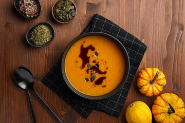 One deep black bowl with an orange pumpkin cream soup with pumpkin seed oil, two black spoons on a wooden surface, top view