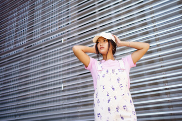 low angle of glad asian female in casual clothes, wearing cap, standing near modern building.