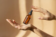 Bottle of serum in women's hands. Glass bottle with dropper cap in women's hands. Amber glass container with dropper lid for cosmetic products on brown background in sunlight
