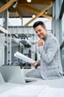 © Westend61 - Smiling businessman holding paper sitting with hand on chin by laptop at desk