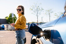 Smiling Woman With Smart Phone Standing By Electric Car At Charging Station