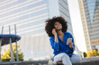 © Westend61 - Smiling woman with Afro hairstyle holding mobile phone sitting on wall