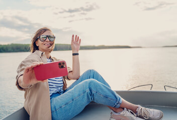 Wall Mural - Young adult caucasian happy woman with smartphone relaxing on a boat on the river enjoying sunset