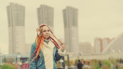 Wall Mural - Young beautiful woman listening to music in wireless headphones on city street. Vivid female with multicoloured hairstyle enjoying songs on walk in cloudy weather.