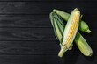 A few corn in leaves lie on a wooden dark background. View from above, space for text