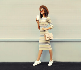 Poster - Portrait of beautiful smiling young woman drinking coffee wearing knitted dress, round hat on city street background