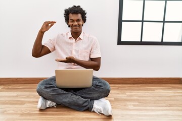 Wall Mural - African man with curly hair using laptop sitting on the floor gesturing with hands showing big and large size sign, measure symbol. smiling looking at the camera. measuring concept.