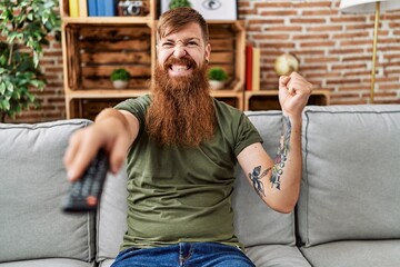 Sticker - Redhead man with long beard holding television remote control screaming proud, celebrating victory and success very excited with raised arm