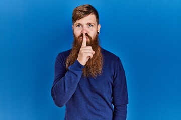 Wall Mural - Redhead man with long beard wearing casual blue sweater over blue background asking to be quiet with finger on lips. silence and secret concept.