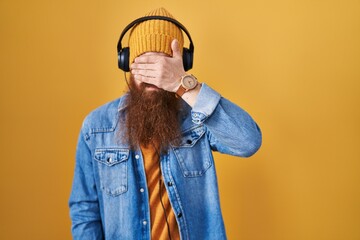 Canvas Print - Caucasian man with long beard listening to music using headphones covering eyes with hand, looking serious and sad. sightless, hiding and rejection concept