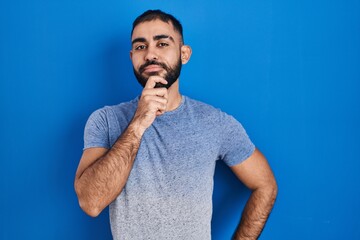 Poster - Middle east man with beard standing over blue background looking confident at the camera smiling with crossed arms and hand raised on chin. thinking positive.