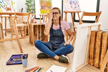 Canvas Print - Young brunette woman at art studio sitting on the floor covering one eye with hand, confident smile on face and surprise emotion.