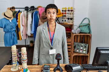 Sticker - Young hispanic woman with short hair working as manager at retail boutique puffing cheeks with funny face. mouth inflated with air, crazy expression.