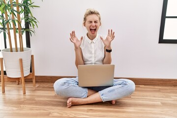 Poster - Young blonde woman using computer laptop sitting on the floor at the living room celebrating mad and crazy for success with arms raised and closed eyes screaming excited. winner concept