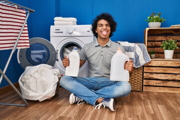 Poster - Hispanic man with curly hair doing laundry holding detergent bottles sticking tongue out happy with funny expression.