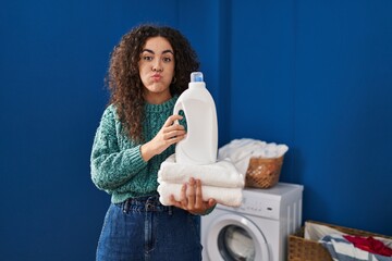 Poster - Young hispanic woman holding laundry and detergent bottle puffing cheeks with funny face. mouth inflated with air, catching air.