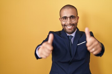 Canvas Print - Hispanic man with beard wearing suit and tie approving doing positive gesture with hand, thumbs up smiling and happy for success. winner gesture.