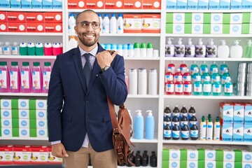 Poster - Hispanic man with beard working as salesman at pharmacy drugstore smiling with happy face looking and pointing to the side with thumb up.