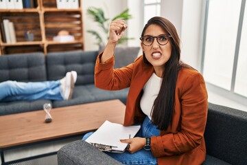 Wall Mural - Young hispanic woman working as psychology counselor angry and mad raising fist frustrated and furious while shouting with anger. rage and aggressive concept.