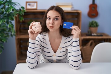 Poster - Young hispanic girl holding piggy bank and credit card puffing cheeks with funny face. mouth inflated with air, catching air.