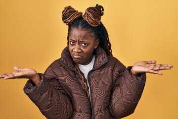 African woman with braided hair standing over yellow background clueless and confused expression with arms and hands raised. doubt concept.