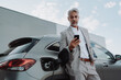 © Halfpoint - Businessman holding smartphone while charging car at electric vehicle charging station, closeup.