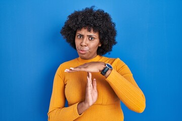 Poster - Black woman with curly hair standing over blue background doing time out gesture with hands, frustrated and serious face