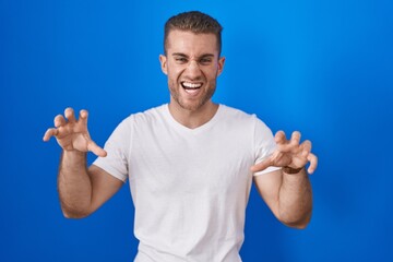 Wall Mural - Young caucasian man standing over blue background smiling funny doing claw gesture as cat, aggressive and sexy expression