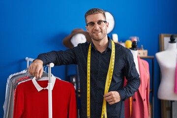 Wall Mural - Young caucasian man tailor smiling confident leaning on clothes rack at clothing factory